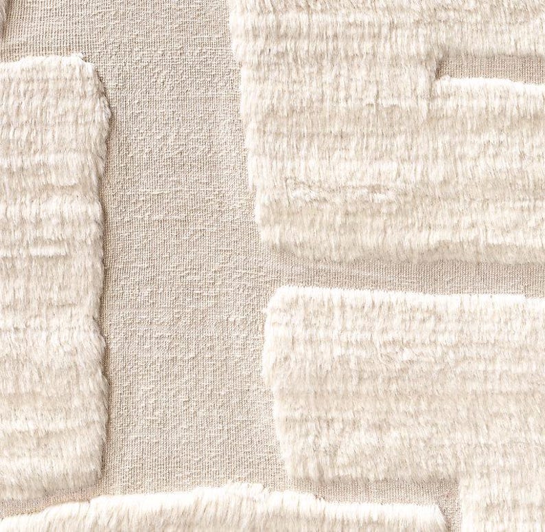 Puede incluir: Primer plano de una tela texturizada de color crema con un dise&ntilde;o geom&eacute;trico. El dise&ntilde;o presenta formas rectangulares en relieve con una textura esponjosa, similar a la piel, creando un contraste t&aacute;ctil con el fondo m&aacute;s liso.