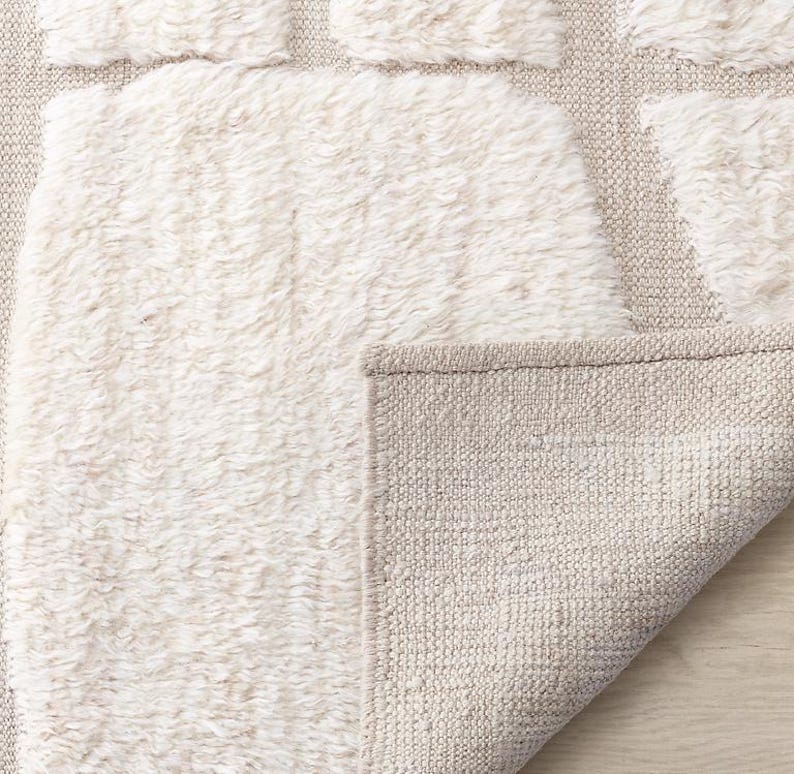 Puede incluir: Una alfombra de tonos neutros con un patr&oacute;n texturizado en blanco roto. El dise&ntilde;o de la alfombra presenta una combinaci&oacute;n de secciones en bucle y tejido plano, creando un efecto en capas. Los bordes est&aacute;n cuidadosamente acabados y una parte del reverso es visible.