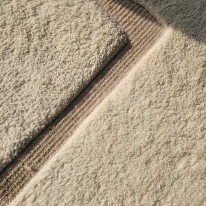 Puede incluir: Primer plano de una alfombra texturizada de color crema con una superficie suave y peluda. Una secci&oacute;n de la alfombra presenta un borde de rayas color canela. El dise&ntilde;o de la alfombra crea una est&eacute;tica acogedora y acogedora, adecuada para la decoraci&oacute;n del hogar.
