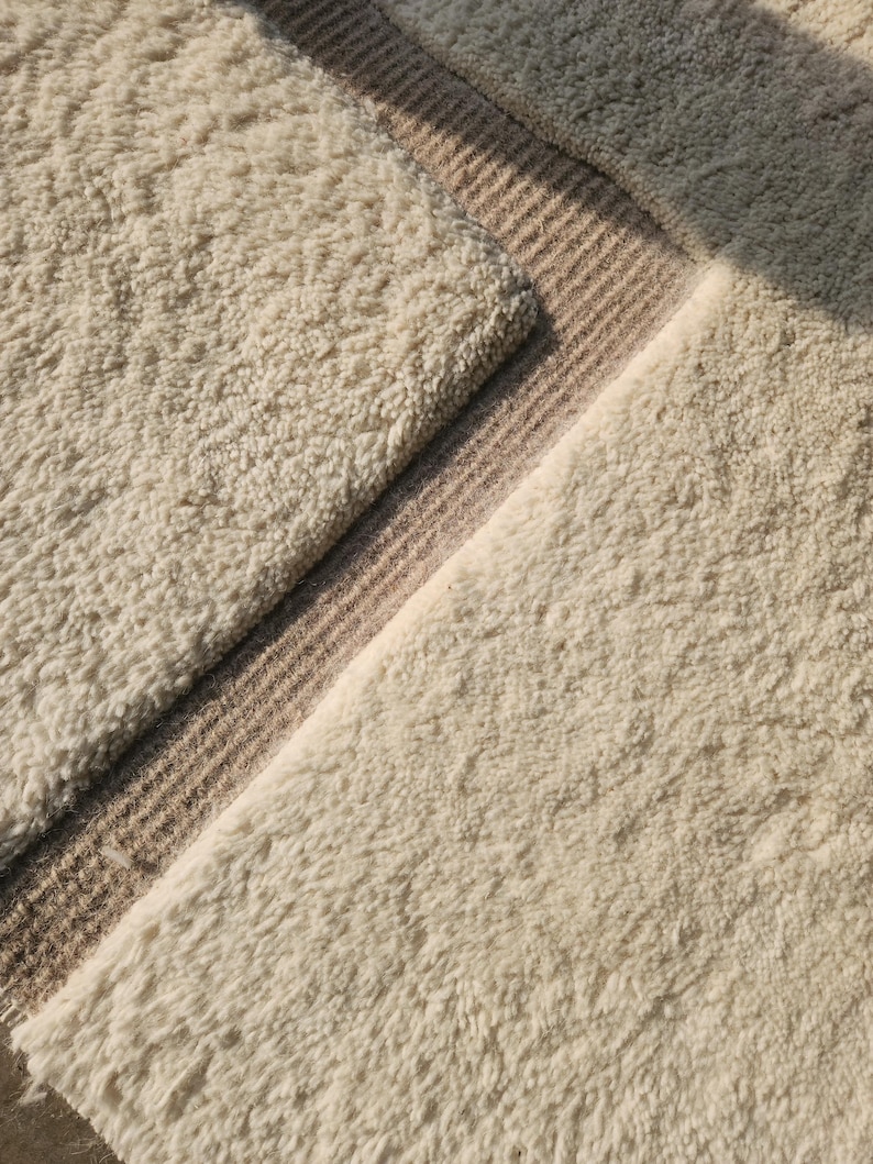 Puede incluir: Primer plano de una alfombra texturizada de color crema con una superficie suave y peluda. Una secci&oacute;n de la alfombra presenta un borde de rayas color canela. El dise&ntilde;o de la alfombra crea una est&eacute;tica acogedora y acogedora, adecuada para la decoraci&oacute;n del hogar.