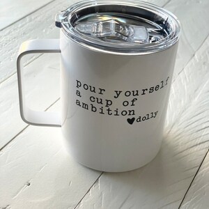 May include: White stainless steel travel mug with a black lid and handle. The mug has the text "pour yourself a cup of ambition &hearts; dolly" printed on the side.
