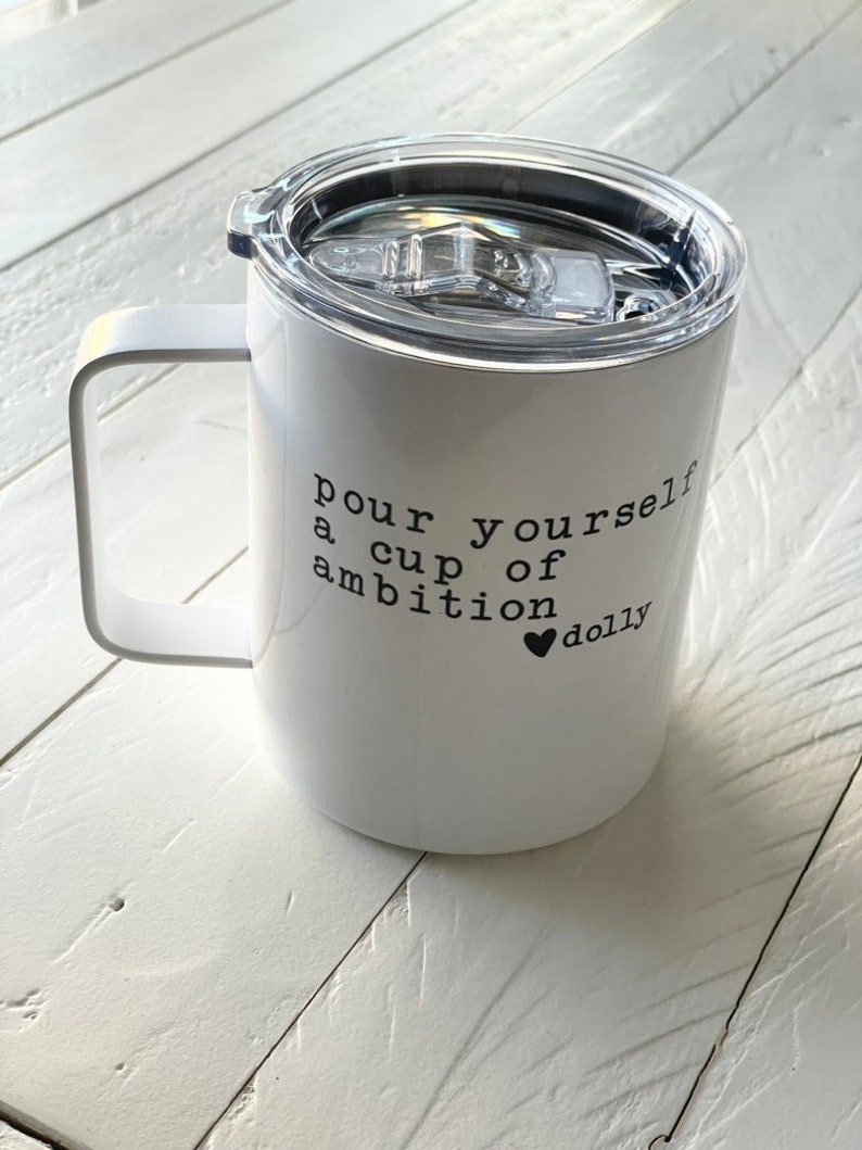 May include: White stainless steel travel mug with a black lid and handle. The mug has the text "pour yourself a cup of ambition &hearts; dolly" printed on the side.