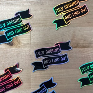 May include: Several black banner stickers with the text "FUCK AROUND AND FIND OUT" in a holographic, iridescent font. The stickers are scattered on a light wood-grain surface.