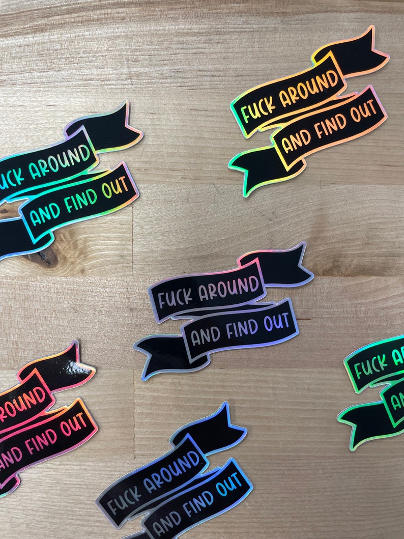 May include: Several black banner stickers with the text "FUCK AROUND AND FIND OUT" in a holographic, iridescent font. The stickers are scattered on a light wood-grain surface.