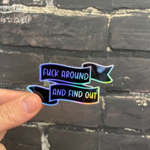 May include: A holographic sticker with the text "FUCK AROUND" above "AND FIND OUT" in a black banner design. The sticker has a shiny, iridescent finish, reflecting various colors. The sticker is held by a hand.