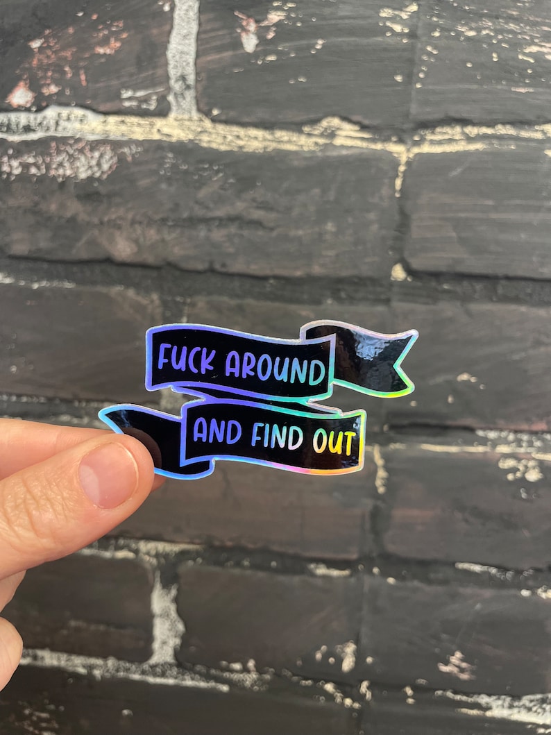 May include: A holographic sticker with the text "FUCK AROUND" above "AND FIND OUT" in a black banner design. The sticker has a shiny, iridescent finish, reflecting various colors. The sticker is held by a hand.