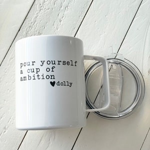 May include: White ceramic mug with a black handle and a black inscription that reads "pour yourself a cup of ambition &hearts; dolly". The mug is sitting on a white wooden surface.