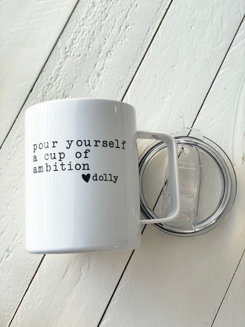May include: White ceramic mug with a black handle and a black inscription that reads "pour yourself a cup of ambition &hearts; dolly". The mug is sitting on a white wooden surface.