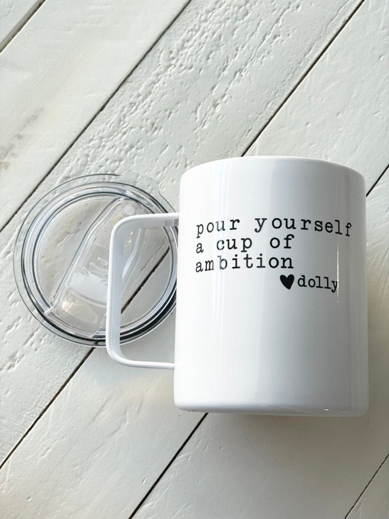 May include: White ceramic mug with a black handle and a black lid. The mug has the text "pour yourself a cup of ambition &hearts; dolly" printed on it.