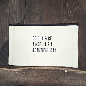 May include: White canvas zippered pouch with black text that reads "Go out & be a hoe. It's a beautiful day."