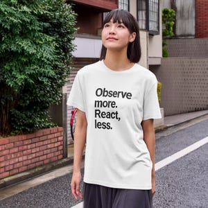 May include: A white t-shirt with the text "Observe more. React, less." in black. The shirt is being worn by a person standing on a street.