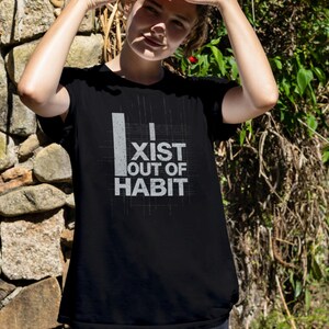 May include: Black t-shirt with the text "I EXIST OUT OF HABIT" in white. The shirt has a simple, modern design. The image shows a person wearing the shirt outdoors.