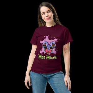 May include: Burgundy t-shirt with a graphic of two cartoon monster heads with purple skin, yellow eyes, and red tongues. The design includes the text "Mad Minds" in a green, gothic font. The shirt is worn by a person.