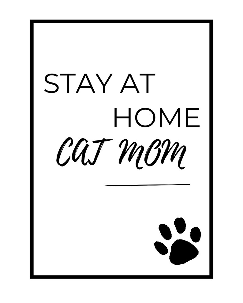 May include: A monochrome print featuring the text "STAY AT HOME cat mom". The words are in a mix of fonts, with a paw print at the bottom right. The print is framed in a black border.