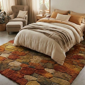 以下が含まれることがあります： ベージュの寝具と茶色の毛布が置かれたベッドがある寝室のインテリア。茶色、オレンジ、黄色の色合いの、石のような模様の大きなテクスチャード加工のラグが床を覆っています。ベージュのカーテンのある窓の近くに椅子とオットマンがあります。