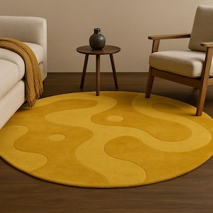 Puede incluir: Una alfombra redonda de color amarillo mostaza con un diseño abstracto y fluido en diferentes tonos de amarillo. La alfombra está colocada sobre un suelo de madera oscura, con un sofá, un sillón y una mesa auxiliar en el fondo. El diámetro de la alfombra es de aproximadamente 183 cm.