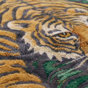 May include: Close-up of a rug featuring a tiger's face. The rug has a textured surface with a design in shades of gold, gray, and green. The tiger's eye is visible, with a detailed pattern of stripes.