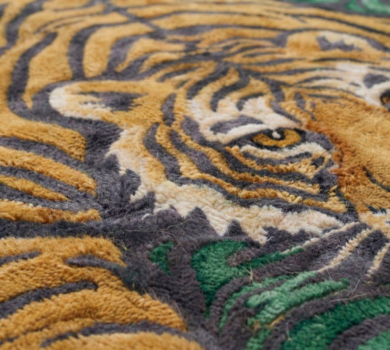 May include: Close-up of a rug featuring a tiger's face. The rug has a textured surface with a design in shades of gold, gray, and green. The tiger's eye is visible, with a detailed pattern of stripes.