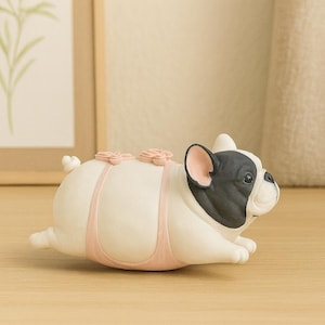 May include: A figurine of a French Bulldog in a lying position. The dog is primarily white with a black patch covering its face and ears. It wears a light pink harness featuring two small, sculpted pink flowers on the back and a small tail.