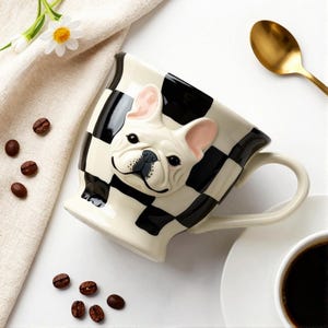 May include: A ceramic mug featuring a black and white checkered pattern and a three-dimensional sculpted French bulldog face on the front. The mug has a curved handle and a glossy finish, showcasing a detailed dog face with pink inner ears.