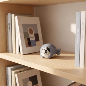 May include: A small, rounded figurine of a French Bulldog wearing a grey seal costume with dark grey spots. The dog has a black and white face with a flat nose and dark eyes. The figurine is positioned on a light wood shelf between books.
