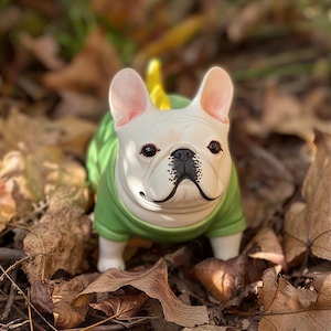 May include: A white French Bulldog figurine with large upright ears, dark eyes, and a black nose, wearing a solid green shirt. A small yellow tail is visible at the back of the figure. The dog is standing in a forward-facing position.