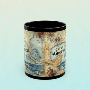 May include: A cylindrical ceramic mug with a black interior and rim. The exterior features a vintage-style map print of the Gulf of America, including illustrations of sailing ships, a compass rose, and the text Gulf of America in black font.