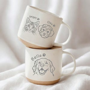 May include: Two stacked ceramic mugs with a cream-colored finish and a light brown base. The top mug features a cat and a dog illustration with the names "Angel" and "Cleo". The bottom mug has a dog illustration and the name "Bella".