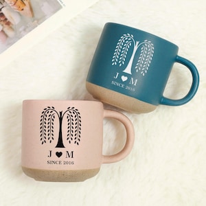 May include: Two ceramic mugs, one teal and one pink, with a tree design and the text "J ♥ M SINCE 2016". The mugs have a light brown base and handle. The mugs are sitting on a white, fluffy surface.