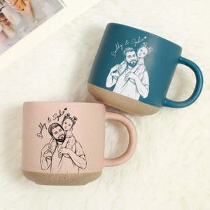 May include: Two ceramic mugs, one teal and one pink, each featuring a sketch of a father carrying a child on his shoulders. The mugs have a speckled texture and a tan base. The text "Daddy & Sophia" is written on each mug.
