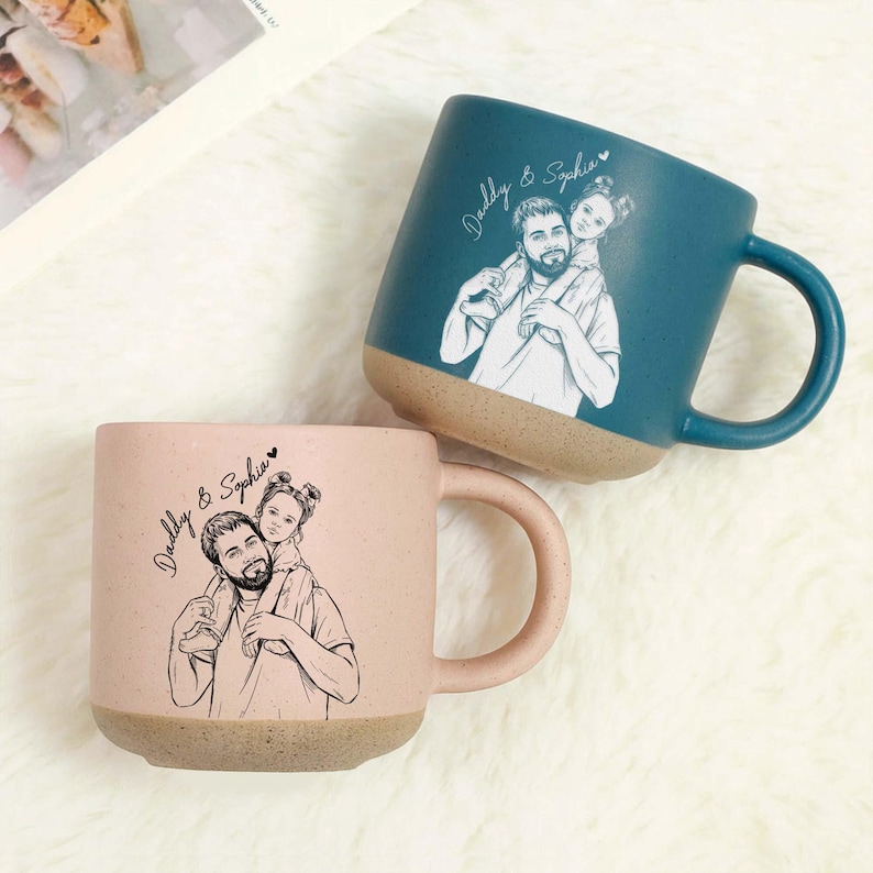 May include: Two ceramic mugs, one teal and one pink, each featuring a sketch of a father carrying a child on his shoulders. The mugs have a speckled texture and a tan base. The text "Daddy & Sophia" is written on each mug.