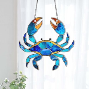 May include: A stained glass crab suncatcher with a blue and gold design. The crab has blue claws with orange tips and is suspended by a silver chain. The crab's body is a mosaic of blue and turquoise glass.