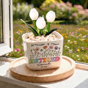 May include: A white ceramic planter with a floral design and the word "RETIRED" in colorful letters. The planter holds glass tulip flowers. The text "Not my problem anymore" and the name "Amanda" are also on the planter.