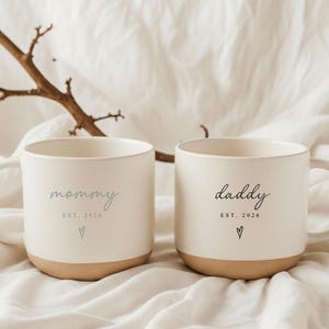 May include: Two off-white ceramic mugs with a beige base. One mug reads "mommy" with "EST. 2026" and a heart. The other mug reads "daddy" with "EST. 2026" and a heart. The mugs are on a white fabric background.