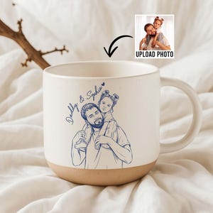 May include: Cream-colored ceramic mug with a brown base and a large handle. The mug features a blue line drawing of a father carrying a child on his shoulders, with the text "Daddy & Sophia". An image of the photo to be uploaded is in the top right.