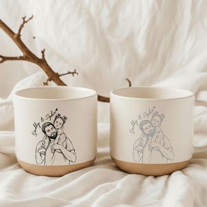 May include: Two ceramic mugs with a cream-colored upper half and a tan base. Each mug features a black and white illustration of a person carrying a child on their shoulders, with the text "Daddy & Sophie" in script.