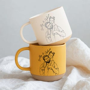 May include: Two ceramic mugs stacked on top of each other. The top mug is off-white, and the bottom mug is yellow. Each mug features a sketch of a father and child, with the text "Daddy & Sophia".