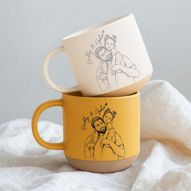 May include: Two ceramic mugs stacked on top of each other. The top mug is off-white, and the bottom mug is yellow. Each mug features a sketch of a father and child, with the text "Daddy & Sophia".