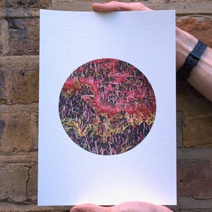 May include: A white paper print featuring a circular design with a textured, abstract pattern. The pattern is predominantly red and pink, with hints of yellow and green, set against a dark background. The print is held against a brick wall.