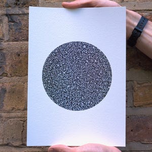 May include: A white rectangular print featuring a large, textured black and gray circle. The artwork is held by two hands against a brick wall background. The circle's pattern appears to be made up of many small, irregular shapes.