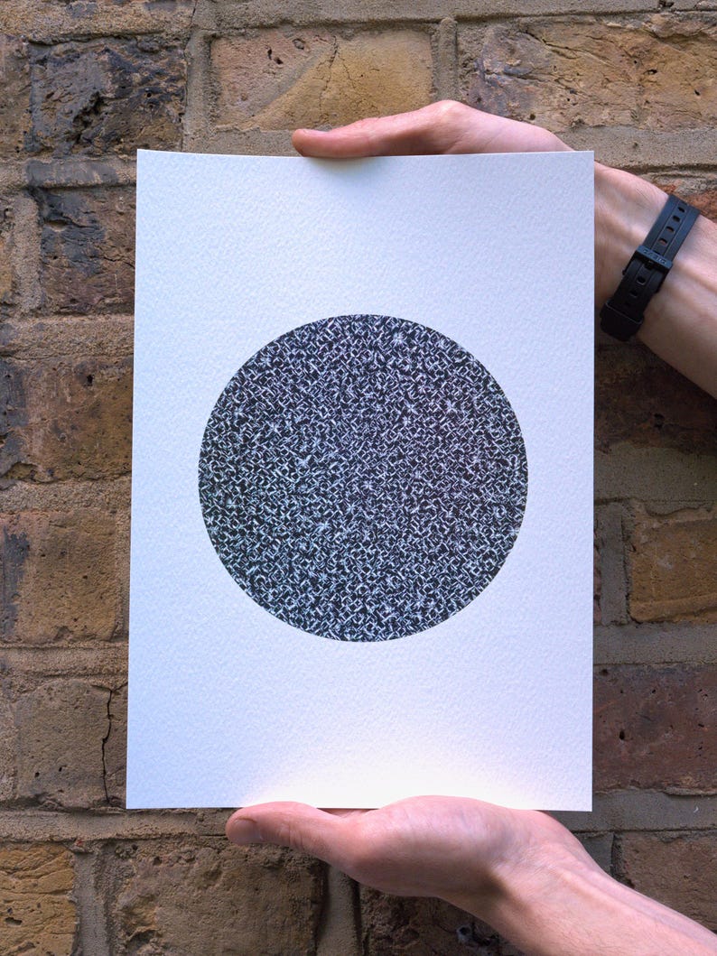 May include: A white rectangular print featuring a large, textured black and gray circle. The artwork is held by two hands against a brick wall background. The circle's pattern appears to be made up of many small, irregular shapes.