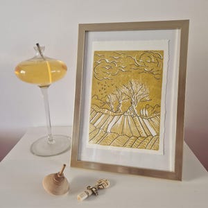 Può includere: Una stampa artistica incorniciata di un paesaggio con tonalità oro e bianco. L'opera raffigura due alberi in un campo sotto un cielo nuvoloso. Sono visibili anche una lampada a olio in vetro, una trottola di legno e un piccolo oggetto decorativo.