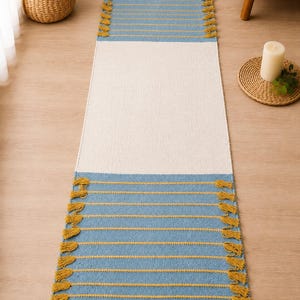 Puede incluir: Una alfombra rectangular en azul y blanco. Las secciones superior e inferior son azules con rayas horizontales amarillas y borlas. La sección central es blanca. Esta alfombra es adecuada para un pasillo o entrada.