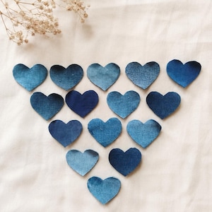 Può includere: Una collezione di quattordici ritagli di tessuto a forma di cuore disposti in una formazione triangolare. Ogni cuore è realizzato in materiale denim, mostrando varie tonalità di blu che vanno dal lavaggio chiaro all'indaco profondo.