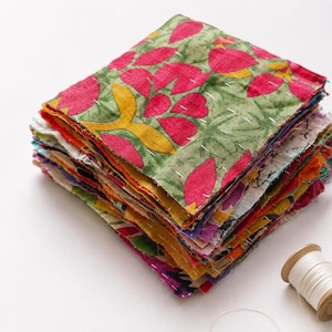 May include: A stack of colorful fabric squares, each with a unique pattern. The top square features a green background with pink floral designs and yellow accents. The edges are stitched with white thread.