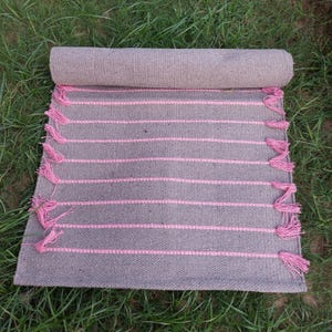 K&ouml;nnte beinhalten: Eine aufgerollte graue Matte mit rosa horizontalen Streifen und rosa Quasten an den R&auml;ndern. Die Matte besteht aus einem gewebten Material und wird auf gr&uuml;nem Gras pr&auml;sentiert.