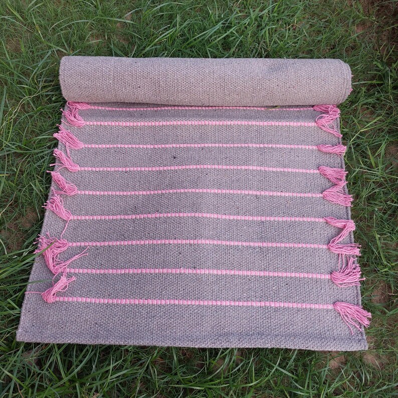 K&ouml;nnte beinhalten: Eine aufgerollte graue Matte mit rosa horizontalen Streifen und rosa Quasten an den R&auml;ndern. Die Matte besteht aus einem gewebten Material und wird auf gr&uuml;nem Gras pr&auml;sentiert.