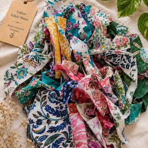 May include: A pile of assorted cotton fabric scraps featuring various floral and botanical patterns in shades of blue, pink, green, and yellow. A tan paper tag is attached, printed with the text Cotton Fabric Waste, Reused. Repurposed. Sustainable.