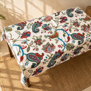 Op de afbeelding: Een rechthoekig wit tafelkleed met een levendig bloemen- en paisley-patroon in rood, blauw en teal. Het tafelkleed bedekt een donkere houten tafel en creëert een decoratief oppervlak voor dineren of presenteren. Ongeveer 150 cm x 200 cm.