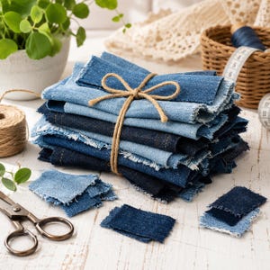 May include: A stack of rectangular denim fabric scraps in various shades of blue, featuring frayed edges and tied together with a brown twine bow. The collection includes several loose, smaller denim pieces scattered nearby on a white surface.